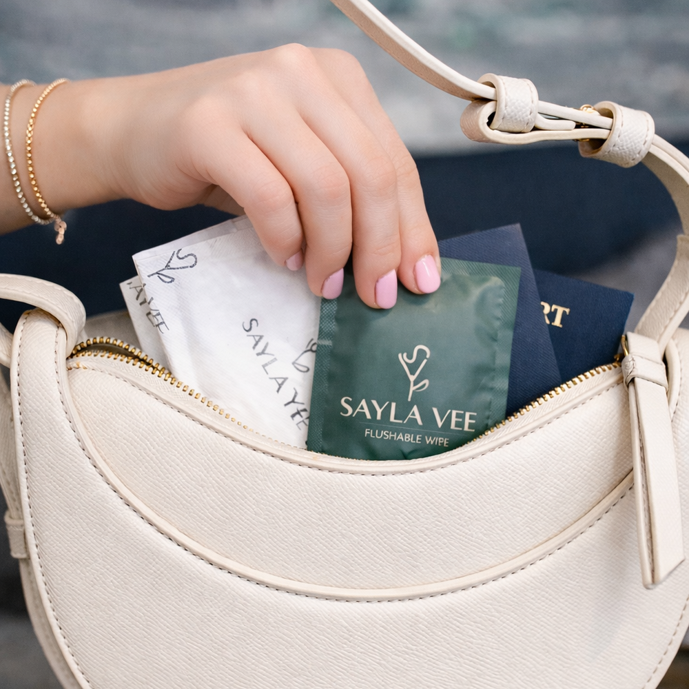 Hand reaching into a beige handbag to take out a individually wrapped wet wipe labeled 'Sayla Vee'.